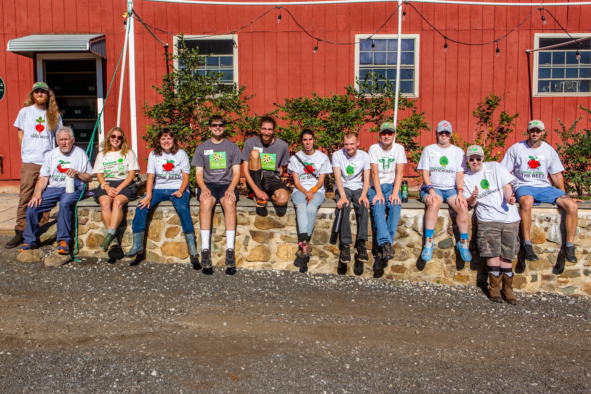 A Farm Less Ordinary growers, staff, and volunteers, pose for a picture in front of a red building. They are all wearing white or greytee shirts with A Farm Less Ordinary branding.